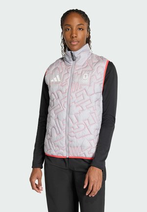 Frau trägt eine hellgraue gesteppte Weste mit roten Akzenten und ein schwarzes Langarmshirt, steht vor einem schlichten Hintergrund und schaut nach vorne.