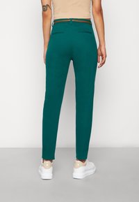 Tyrkysové skinny kalhoty s hladkým povrchem, zadními kapsami a hnědým opaskem; stylizované s bílými teniskami s béžovým akcentem.