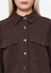 Brun denimskjorte med krage, sølvfargede knapper og to brystlommer med klaff. Glatt tekstur og tilpasset passform.
