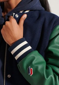 Persoon draagt een marineblauw en groen varsityjack, houdt de kraag vast met één hand; het jack heeft gestreepte manchetten en een klein rood-wit-blauw logo.