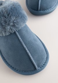 Blaue Wildlederschlupfschuhe mit flauschigem Pomponbesatz und sichtbarer Naht auf hellem Hintergrund.