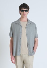 Camisa de botones de manga corta, gris claro, con una textura sutil, combinada con una camiseta de cuello redondo beige y pantalones de color claro.