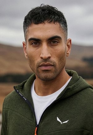 Mand med kort krøllet hår iført en grøn lynlåstrøje over en hvid skjorte, stående udendørs med slørede bakker i baggrunden.