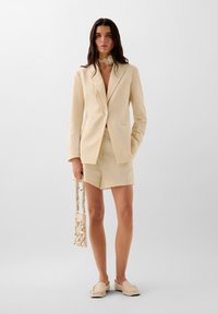 Beige bleiseri, jossa on yksi nappi, yhdistettynä samansävyisiin shorts'eihin. Asusteena helmillä varustettu käsilaukku ja liukuva kengät.