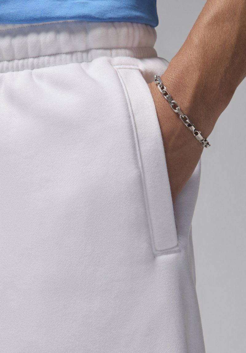 Weiße Jogginghose mit seitlichem Taschen, kombiniert mit einem blauen Hemd. Ein silbernes Armband ist an einem gebräunten Handgelenk sichtbar. Weicher Stoff mit angenehmer Textur.