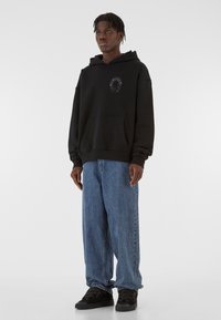 Hanorac negru cu glugă, prevăzut cu o buzunăraș frontal și detalii cu logo. Asortat cu blugi denim bleu, cu croială lejera și adidași negri.