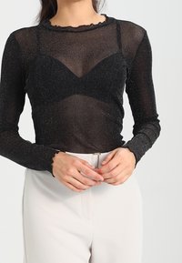 Genomskinlig svart långärmad topp med glittrande textur, volangkrage och figurnära design, lager på en svart bh. Lätt tyg, figursydd form.