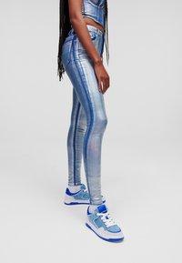 Vartalonsyv ät farkut metallisin sinisin värein, heijastavalla pinnalla ja pystysuorilla raidoilla, yhdistettynä sinisiin ja valkoisiin tennareihin, joissa on denimkorostuksia.