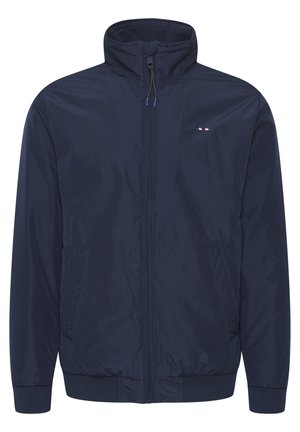 Tummansininen tuulitakki, jossa on korkea kaulus, etuvetoketju, kaksi sivutaskua ja ribbireunat. Takissa on pieni monivärinen logo rinnassa.