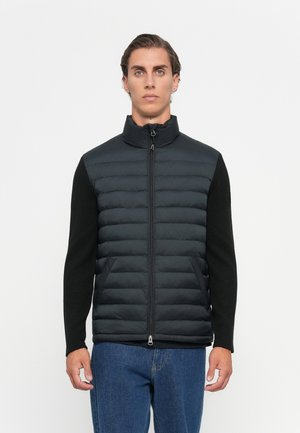 Zwarte puffer vest met een hoge kraag, ritssluiting aan de voorkant en gebreide zwarte mouwen. Twee zijzakken en een gladde textuur. Draag over blauwe jeans.