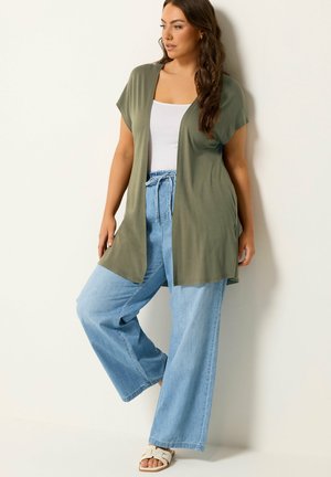 Kvinde iført olivengrøn åben cardigan, hvid tanktop, lyseblå viddejeans og hvide slidesandaler, stående op ad en hvid væg.