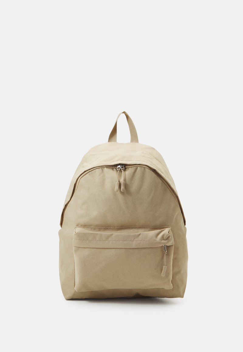Mochila de tela beige con forma redondeada, compartimento principal con cremallera, bolsillo frontal con cremallera y un asa de transporte en la parte superior.
