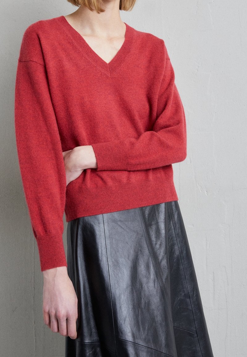 Kvinna klädd i en röd V-ringad tröja med långa ärmar och en svart läskjol, stående mot en enfärgad ljusgrå vägg.