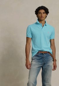 Tânăr cu păr creț, purtând un tricou polo bleumarin deschis, o curea împletită maro și blugi blue jeans decolorati, stând în fața unui fundal neutru.