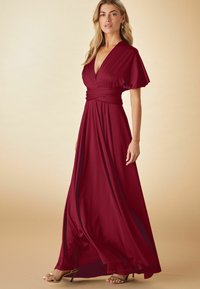Duga, burgundska haljina s dubokim V-izrezom, lepršavim rukavima i uskim strukom. Tkanina koja teče elegantno pada do poda. Zlati sandale s trakama.