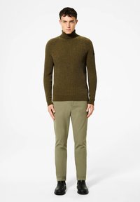 Uomo in piedi che indossa un maglione a collo alto a coste verde oliva, pantaloni verde oliva chiaro e scarpe nere su uno sfondo bianco semplice.