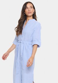 Gestreiftes Kleid in Blau und Weiß mit Knopfleiste, aufgerollten Ärmeln und einer gebundenen Taille. Die Länge reicht bis zur Mitte der Wade mit seitlichen Schlitzen.