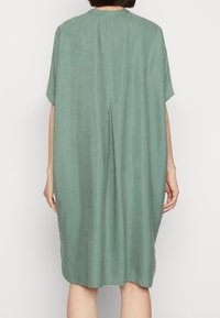 Zelené šaty po kolena vyrobené z jemné tkaniny. Mají krátké rukávy, kulatý výstřih a šev uprostřed zad s malým skladem.