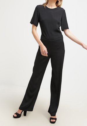 Femme portant un haut noir ajusté à manches courtes et un pantalon noir taille haute à jambes larges, avec des sandales noires à talons hauts.