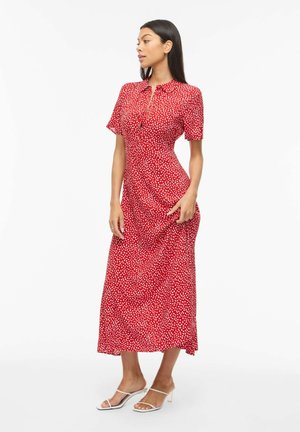 Frau trägt ein rotes, kurzärmeliges Midikleid mit weißen Punkten und Knöpfen, steht seitlich und hält das Kleid mit einer Hand.