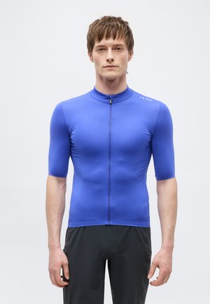 Férfi szemben áll, kék, rövid ujjú, testhezálló kerékpáros mezben és fekete nadrágban.