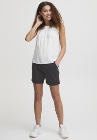 Hvit ermeløs topp med folder, kombinert med mørkegrå rullede shorts. Modellen har på seg hvite sneakers og poser med en hånd på nakken.