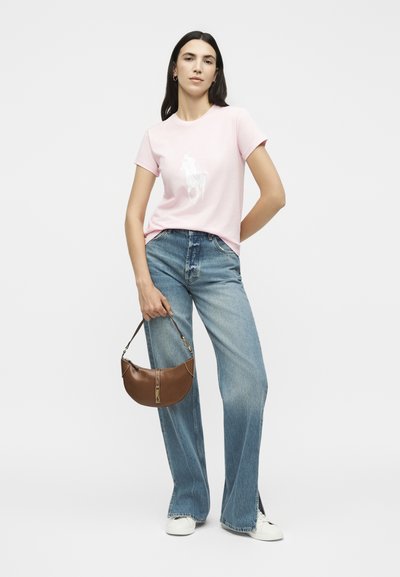 Γυναίκα που στέκεται, φορόντας ένα ανοιχτό ροζ μπλουζάκι με λευκό γραφικό άλογο, φαρδιά μπλε τζιν, λευκά αθλητικά παπούτσια και κρατώντας μια καφέ δερμάτινη τσάντα.