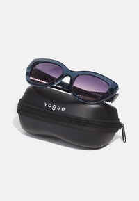 Mėlynos plastikinės saulės akiniai su purpurinėmis gradientinėmis lęšiais ilsisi ant juodo užtrauktuku užsegamo dėklo, pažymėto "vogue" prekės ženklu. Dėklas turi tekstūruotą paviršių.