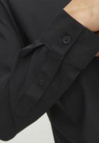 Camicia nera con maniche lunghe, polsini con un solo bottone, tessuto morbido e dettagli di cucitura precisi. Il polsino presenta un accento appuntito.