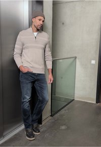 Světlý béžový svetr na zip s horizontálními pruhy, doplněný modrými džínami a pevnými černými botami, na pozadí moderního betonového povrchu.