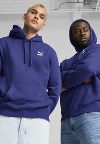 Due individui indossano felpe con cappuccio blu navy abbinate, realizzate in morbido cotone. Ognuna presenta un logo Puma bianco sul petto e una tasca a marsupio.