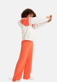 White hoodie with orange sleeves and red stripe details, paired with bright orange wide-leg pants and white sneakers. Smooth fabric texture.