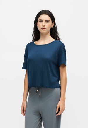Vrouw met lang, krullend donker haar draagt een blauw gebreid kort mouwtopje en losse grijze broek, staand tegen een effen lichte achtergrond.