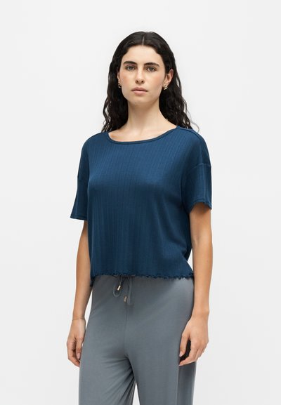 Femme aux longs cheveux bouclés et foncés porte un top en maille bleu à manches courtes et un pantalon gris ample, debout contre un fond clair uni.