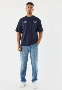 Man staat met handen gedeeltelijk in de zakken, draagt een marineblauw graphic t-shirt, lichtblauwe jeans met patroon en witte sneakers op een witte achtergrond.
