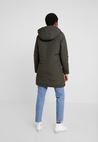 Persoon in een donkergroene gewatteerde parka met capuchon, lichtblauwe spijkerbroek en witte hoge sneakers, staande met de rug naar de camera op een effen achtergrond.