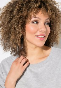 Felpa grigia con scollatura rilassata, indossata da un individuo con capelli ricci. Tessuto leggermente texturizzato visibile, con una mano vicino al colletto.