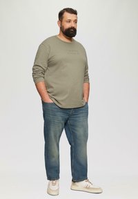 Graues Langarm-T-Shirt mit geprägtem Text, kombiniert mit blauen Jeans und beigen Sneakers. Das Shirt hat einen lockeren Schnitt und eine weiche Textur.