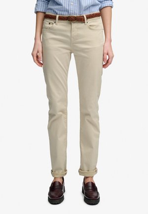 Osoba nosząca beżowe podwinięte spodnie z plecionym brązowym paskiem, niebieską paskowaną koszulę i ciemnobrązowe mokasyny, stojąca na białym tle.