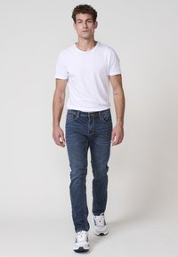 Jeans azules entallados para hombre y una camiseta blanca lisa. Los jeans tienen un diseño de cinco bolsillos y un ligero desgaste, combinados con zapatillas blancas.