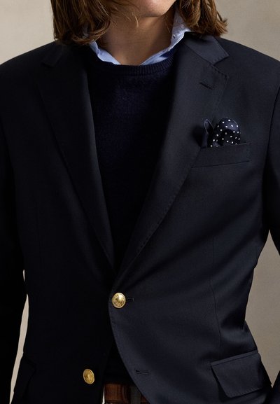 Person iført en mørk marineblazer med guldknapper, en navyblå sweater, lyseblå skjortekrave og et navyblåt lommetørklæde med prikker.