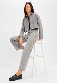 Szara, krótka kurtka z denimu z dwiema kieszeniami na piersi oraz szerokimi spodniami w tym samym kolorze. Czarne platformowe mokasyny dopełniają całość. Gładkie tło.