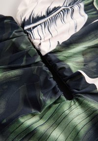 Gros plan sur un tissu vert et blanc avec une fermeture éclair noire et des coutures élastiques froncées formant un motif botanique feuillu.