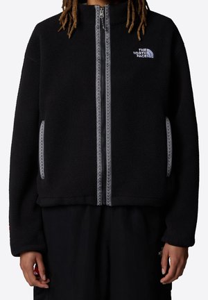 Person trägt eine schwarze Fleecejacke mit Reißverschluss, auf der Brust ist das The North Face-Logo und entlang des Reißverschlusses sowie der Taschen reflektierende Besätze.