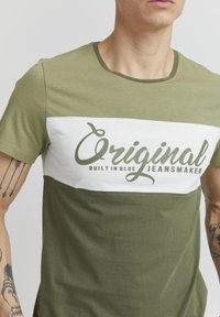 T-shirt kratkih rukava maslinaste zelene boje s bijelim horizontalnim panelom. Na njemu se nalazi veliki grafički natpis tamnozelene boje koji glasi "Original Built In Blue Jeansmaker."