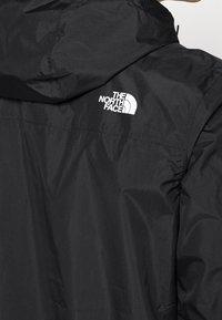 Czarna, lekka kurtka z kapturem, o gładkiej fakturze, z białym logo The North Face na plecach.