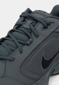 Egy sötétszürke Nike Air Monarch sportcipő közeli felvétele, amelyen a részletes fűzés, a texturált szövet, a varrott átfedések és a fekete Nike pipa logó látható.