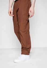 Person som bär bruna cargo-byxor och vita Nike-sneakers, stående mot en enfärgad ljus bakgrund.