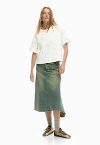 Mujer con una camiseta blanca grande con detalles florales, una falda midi de mezclilla deslavada de talle alto, calcetines grises y zapatillas beige.