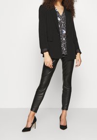 Černý ušitý blazer s klopami, kombinovaný s vzorovanou blůzou s modrými a bílými prvky a černými kalhotami z umělé kůže.